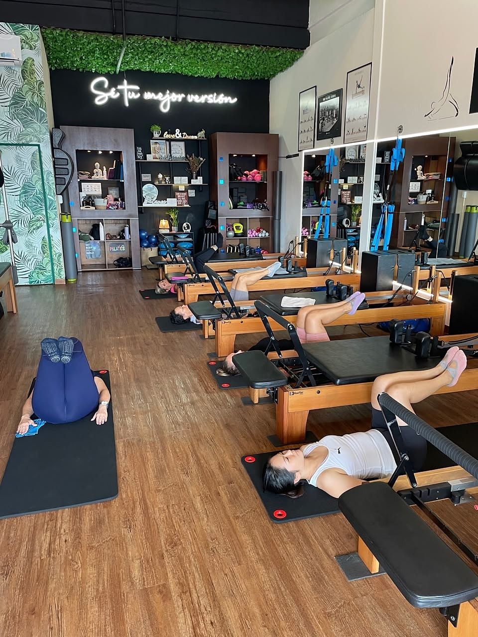 Clase de Pilates Mat: alumnas tumbadas en el suelo y en el Reformer con las rodillas recogidas, frente al letrero «Sé tu mejor versión»