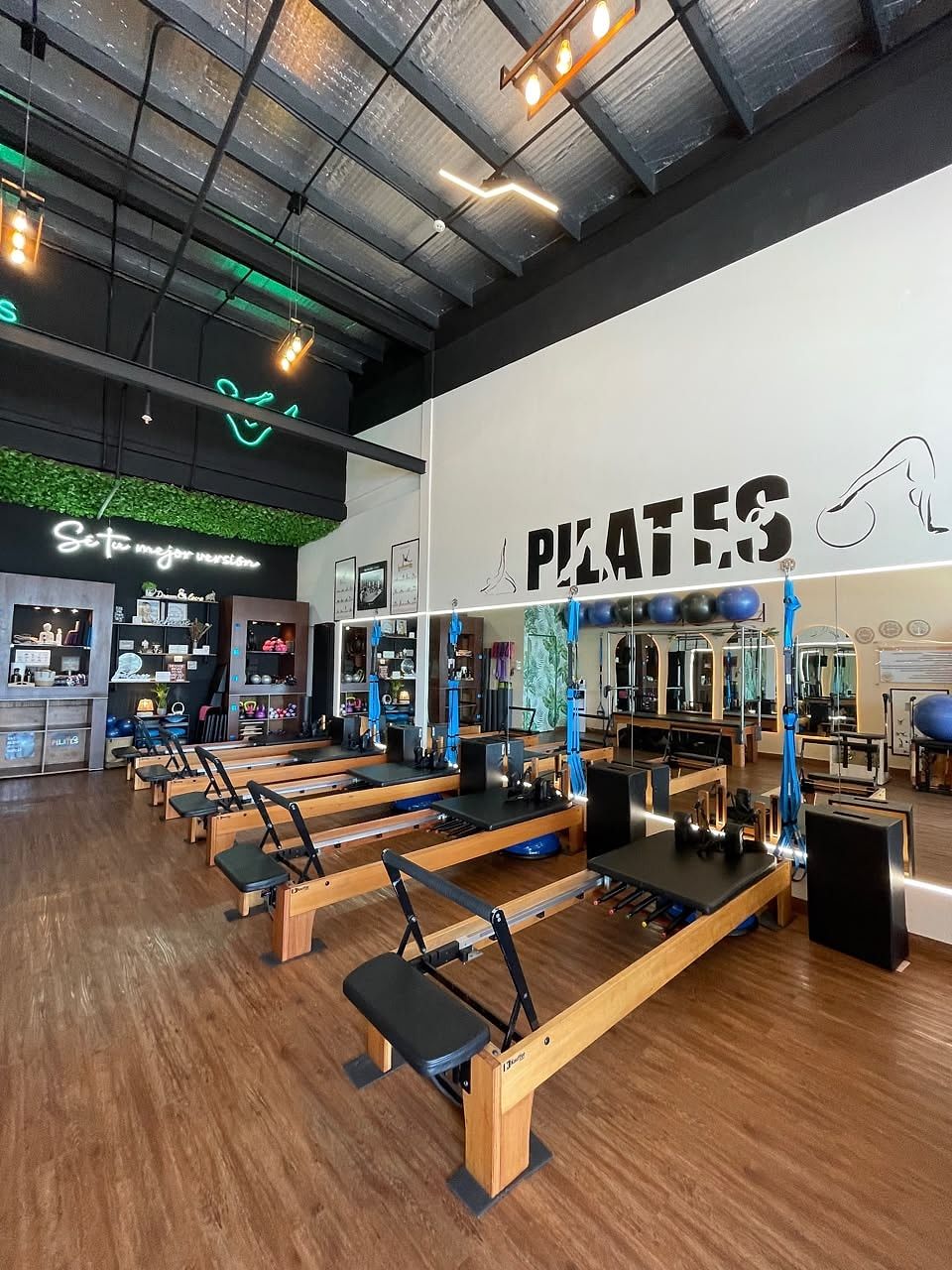 Vista del estudio: fila de máquinas Reformer de madera, techo alto, letrero de neón «Sé tu mejor versión» y la palabra PILATES en la pared